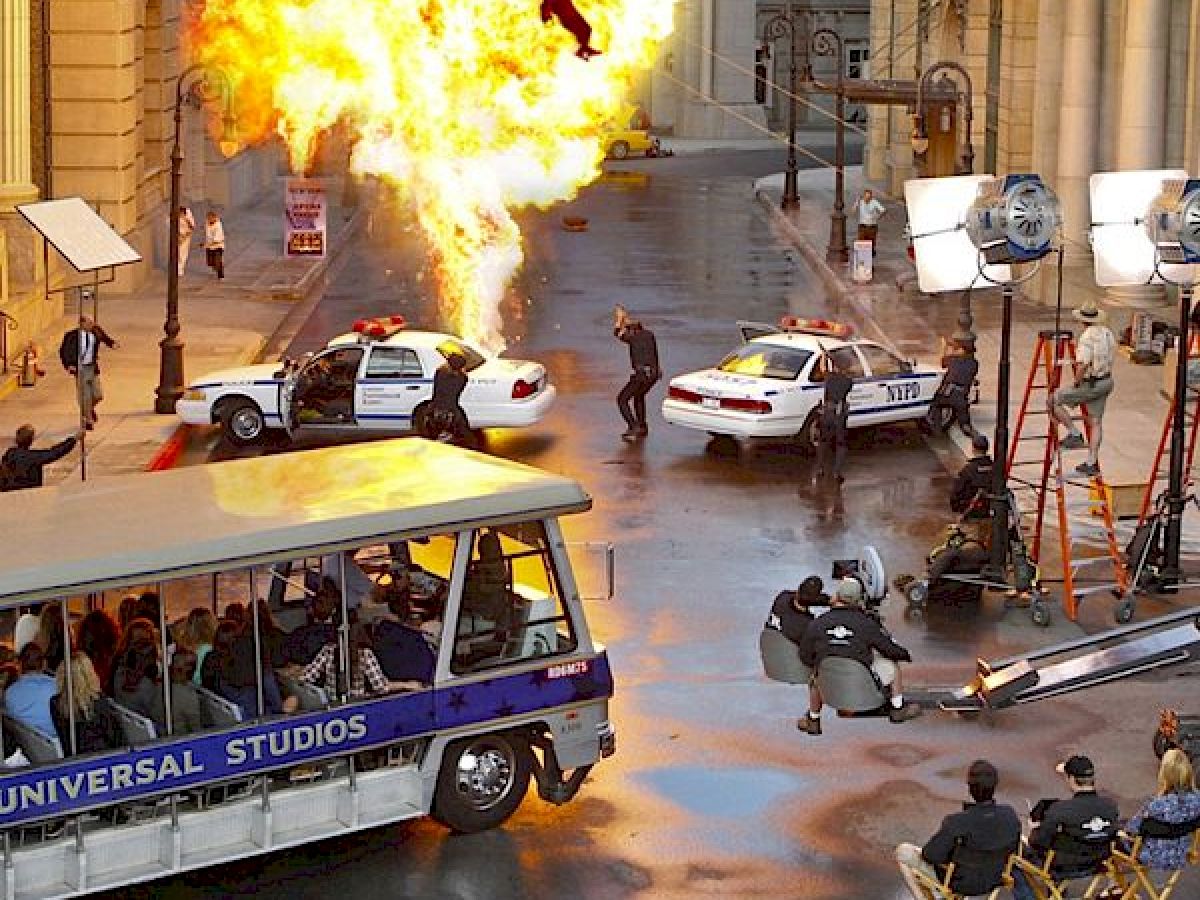 This image shows a movie set with an explosion scene being filmed. There are police cars, film crew, cameras, and a Universal Studios tram.