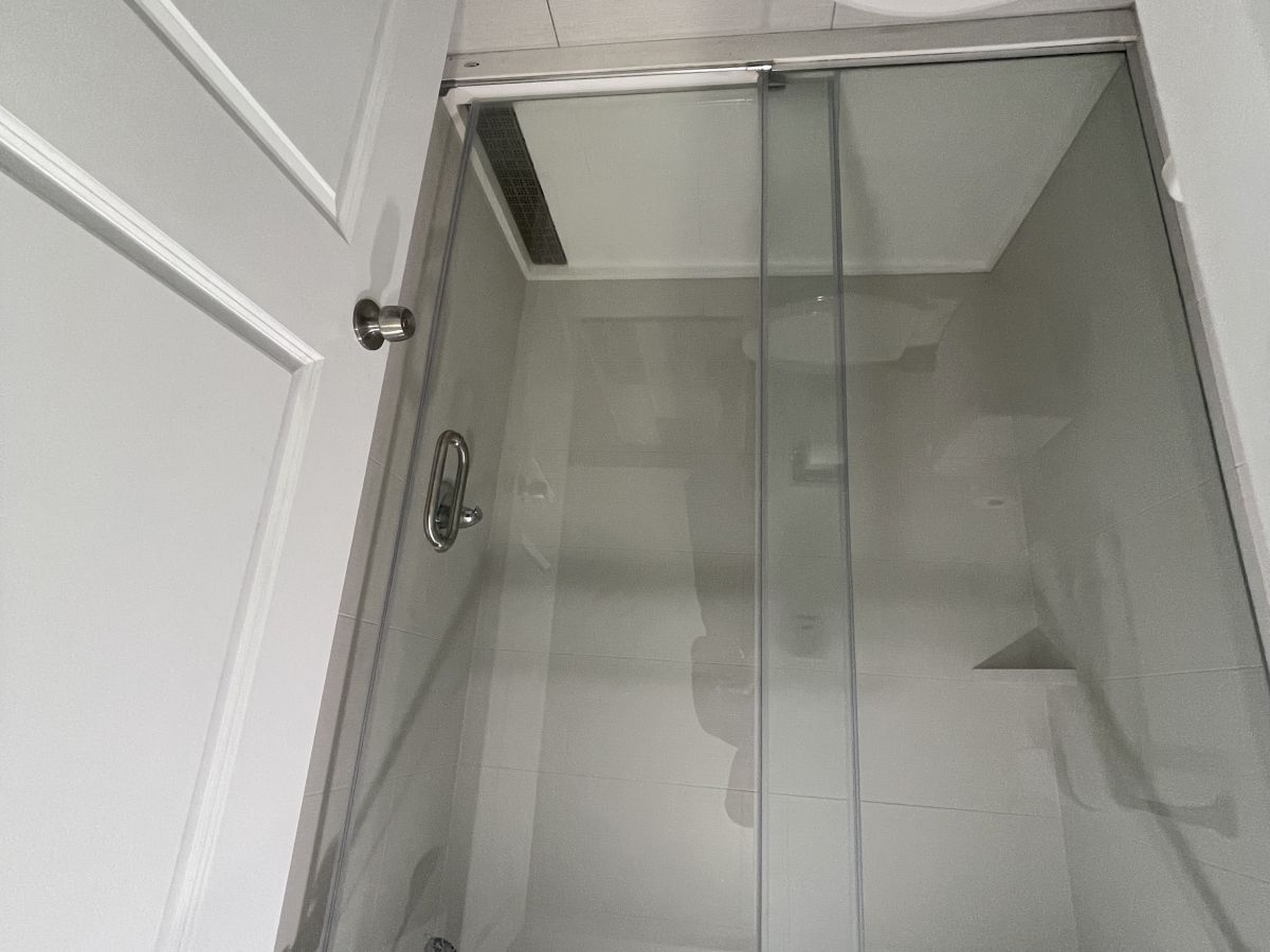 The image shows a bathroom with a glass shower enclosure, a white door, and part of a toilet visible, emphasizing a clean and modern design.