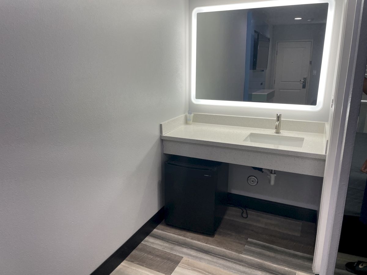 The image shows a modern bathroom with a countertop sink, a lighted mirror, and a dark cabinet below the sink on a laminate floor.