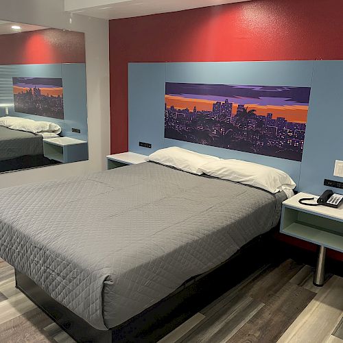 A hotel room features a bed with gray bedding, cityscape artwork, bedside tables, a phone, mirror, and a green chair.