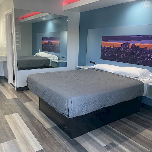 A modern hotel room with a large bed, gray bedding, a cityscape painting above the headboard, a mirror, and a vanity area with a sink and mirror.